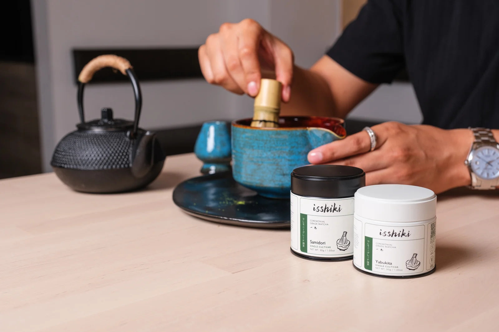 Hands whisking matcha beside Isshiki tins.