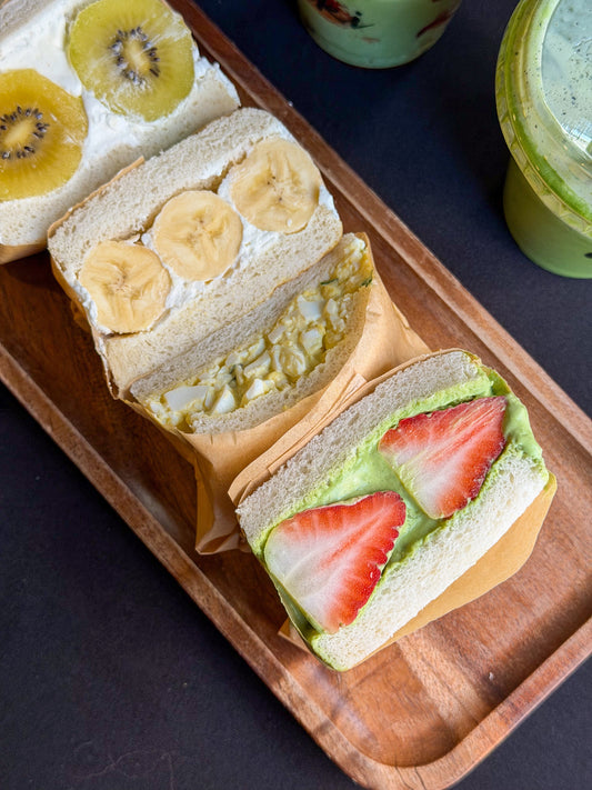 A wooden tray displaying Japanese sandos including strawberry matcha cream, banana cream, and egg salad, served alongside iced matcha drinks, highlighting the Isshiki Matcha café menu and NYC matcha experience.