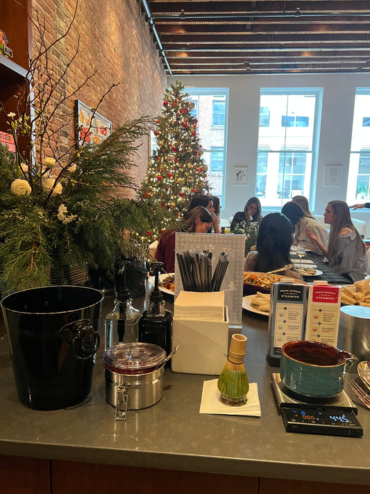 A matcha bar setup with ceremonial matcha tools, whisk, syrup bottles, and drink station in front of guests gathered around a decorated Christmas tree, highlighting Isshiki Matcha event catering at a festive holiday party.