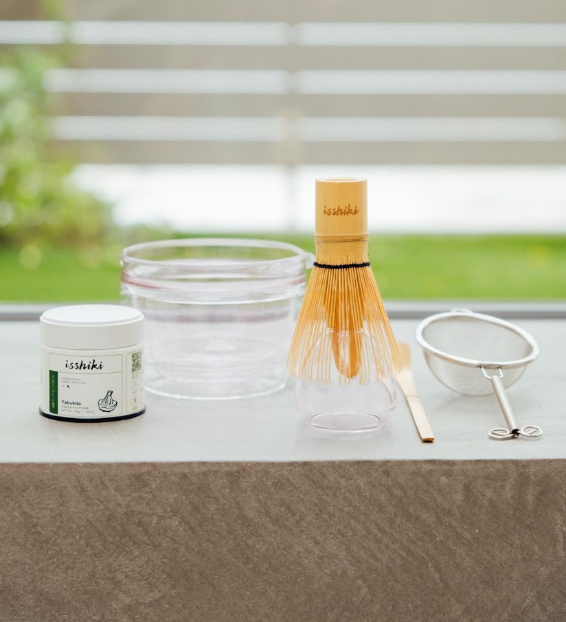 Isshiki x Hario matcha set with glass bowl, bamboo whisk, tin, and strainer arranged on a table by a window