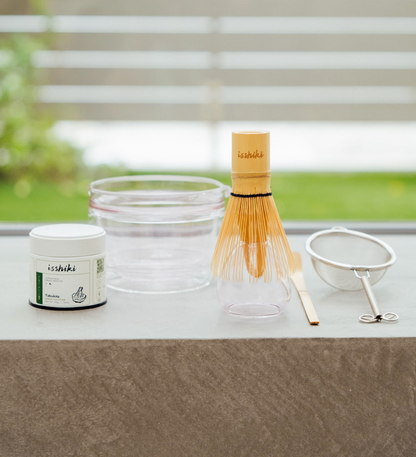 Isshiki x Hario matcha set with glass bowl, bamboo whisk, tin, and strainer arranged on a table by a window