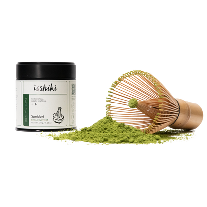 Isshiki Samidori ceremonial matcha tin beside a bamboo whisk resting on a mound of green matcha powder